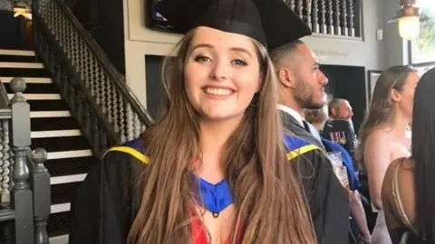 Lucie Blackman Trust Grace Millane pictured at the bottom of a staircase. She is wearing a mortar board hat and black gown for a graduation ceremony. She has long brown hair and is smiling.