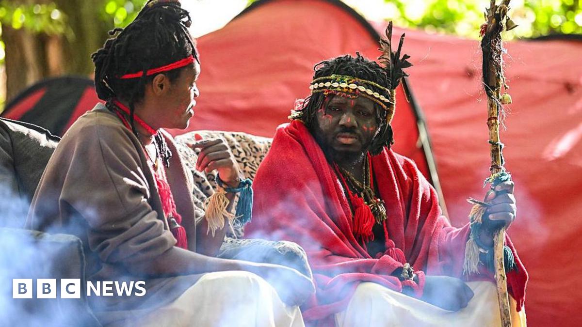 A dark-haired woman with a brown top  and red head band looks towards a man with a red top, facial paints and beaded head bands. Both are seated in front of a red tent