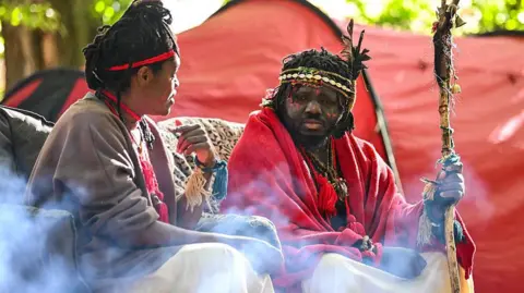 Getty Images A dark-haired woman with a brown top  and red head band looks towards a man with a red top, facial paints and beaded head bands. Both are seated in front of a red tent
