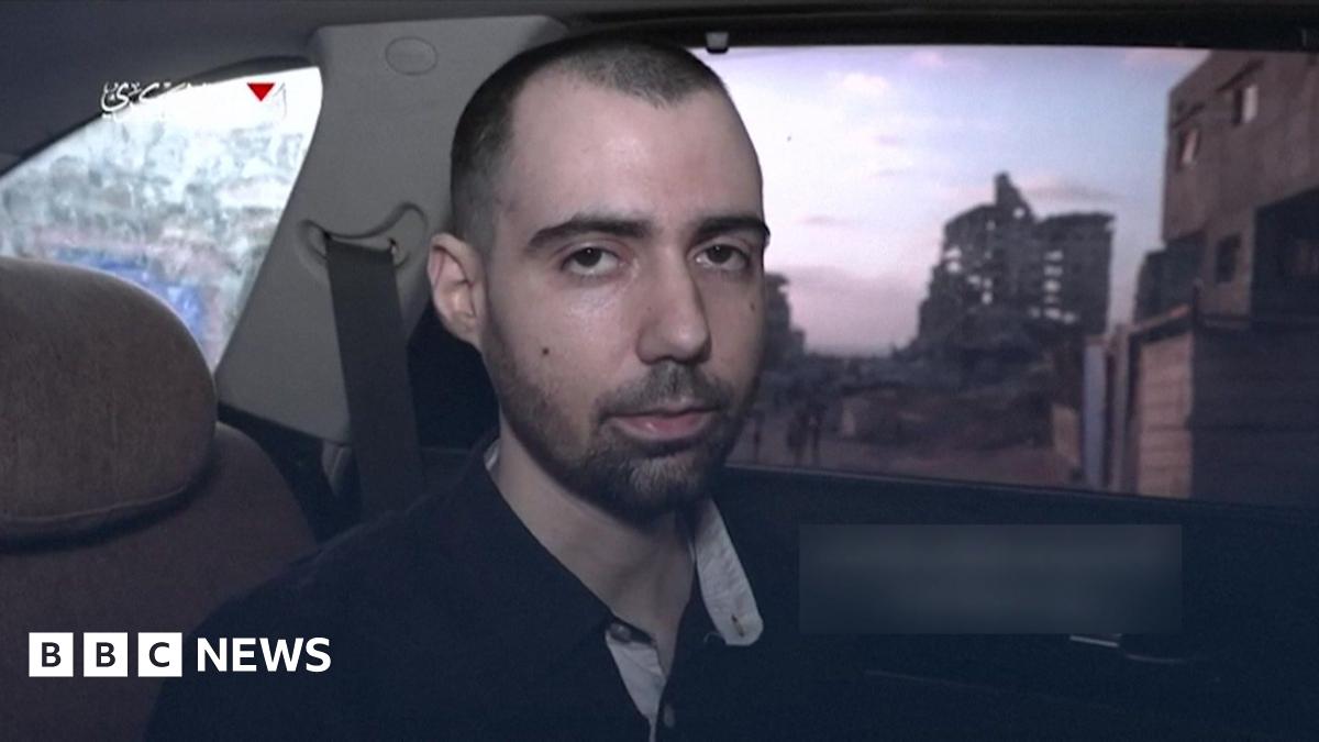 A screenshot of a video released by Hamas, showing Guy Gilboa-Dalal in the back of a car. The picture shows his head and shoulders, as he faces the camera. He has short-cropped dark hair and is wearing a dark-coloured polo shirt. Behind him through a window two buildings can be seen in the background, one of which appears to be damaged.