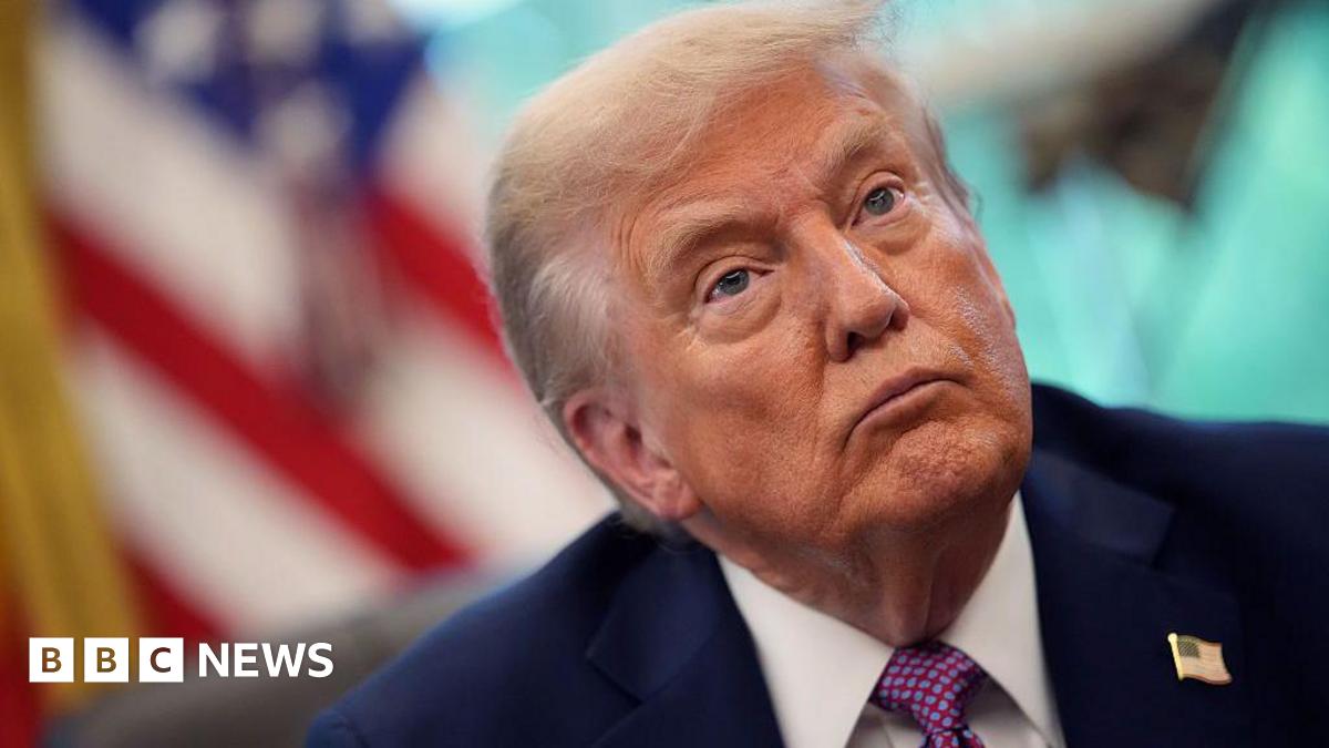 A close up of US President Donald Trump who is wearing a dark blue suit, white shirt and purple tie with blue dots. There is also an American pin on his right lapel.