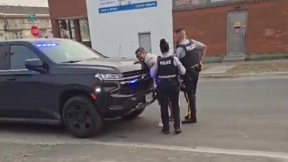 A man being handcuffed by two RCMP officers as he leans against an unmarked police cruiser.