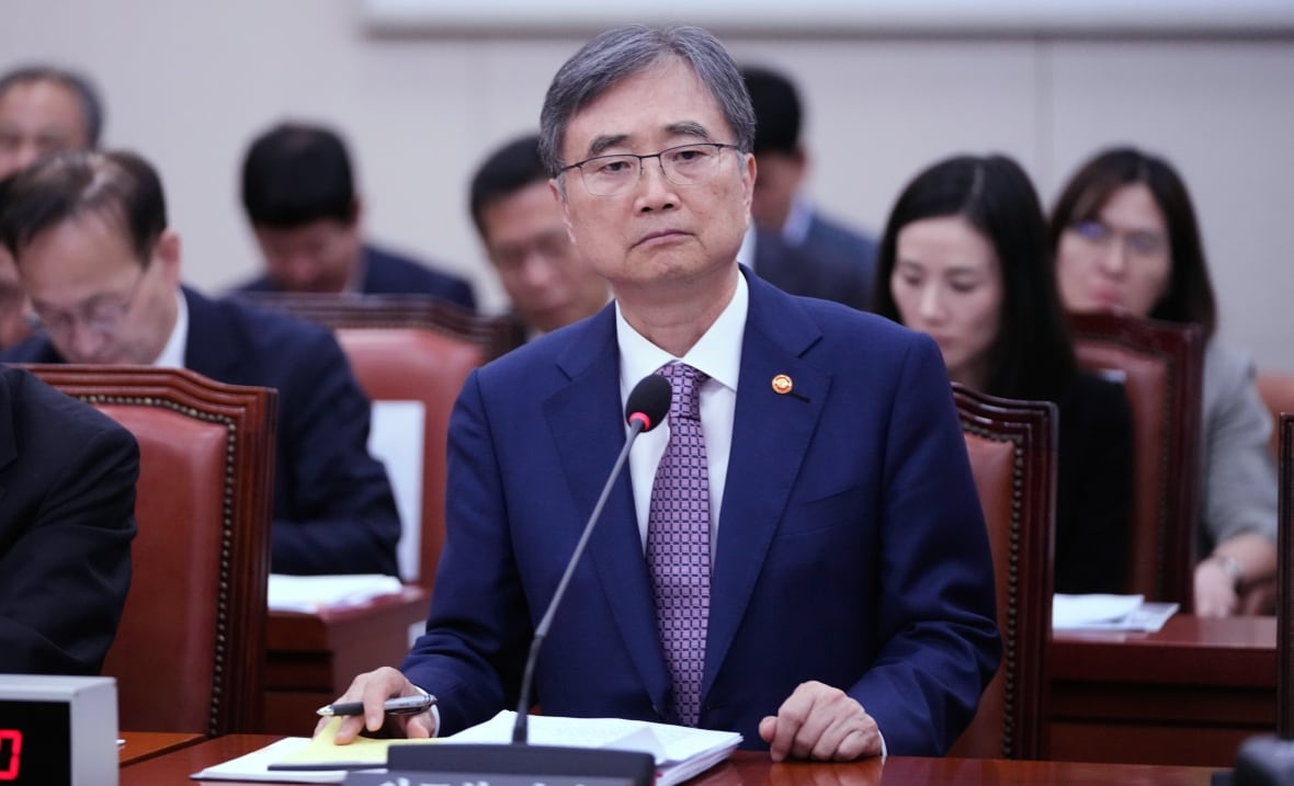 A man in a suit sits at a long desk with a microphone in front of him. Another person sits next to him, and rows of long desks with other people sat at them are visible behind him. 