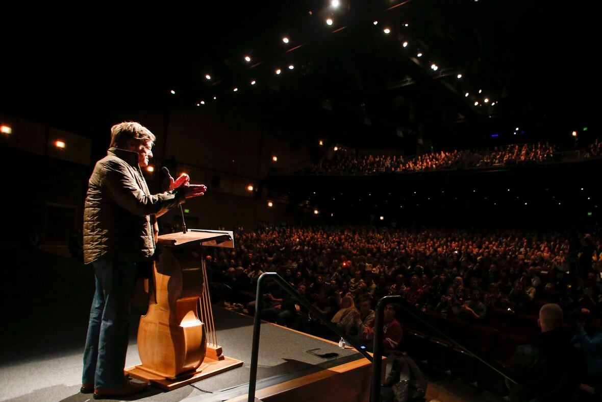 a man in jeans and a plain jacket stands on a stage at a podium. the audience in front of him claps