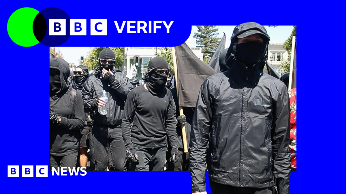 A group of people dressed in all-black, with their eyes and mouths covered by sunglasses and masks, march forward during an anti-fascist protest