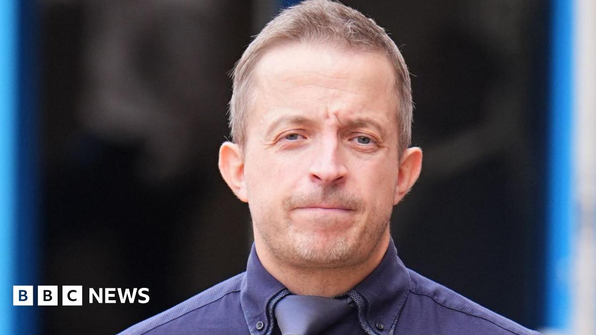 Haden Pentecost wearing a navy blue shirt and tie, looks at the camera