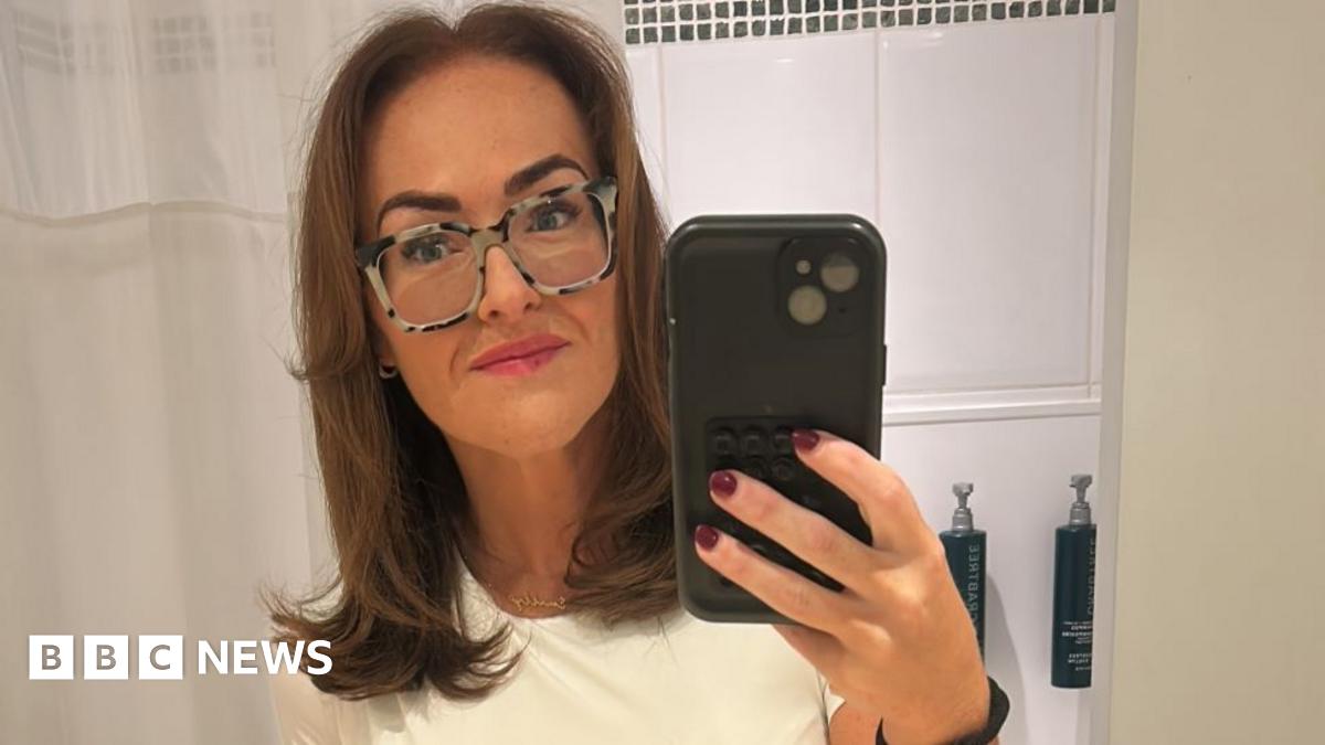 A woman with straight brown hair, a white t-shirt, patterned glasses and pink lipstick takes a selfie in the mirror with a phone