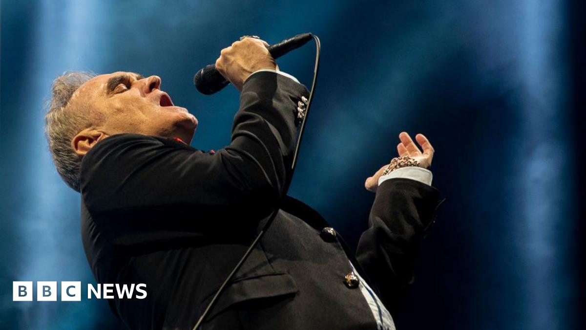 Morrissey bends back as he sings into a microphone, wearing a black suit and tie with a white shirt.