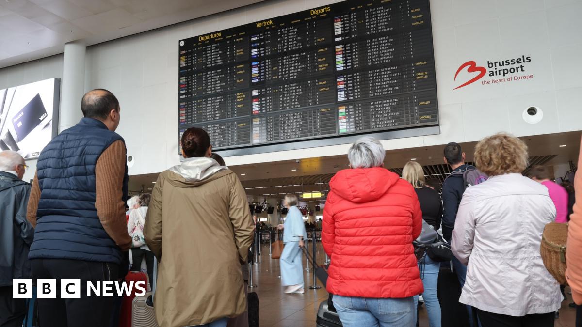 A group of people leaning on wheeled suitcases look up at an airport departures board. In front of them are a series of airport check-in desks.