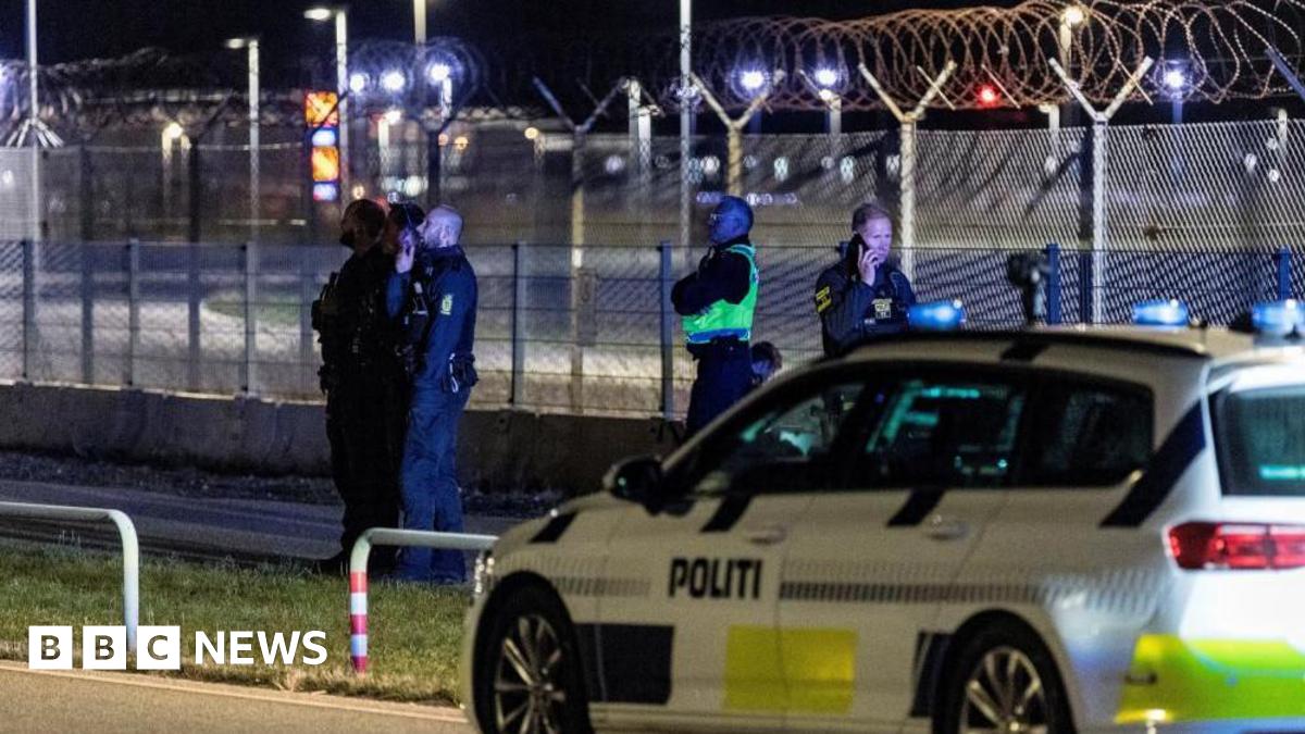Police stand next to a fence outside an airport at night, a police vehicle is in the foreground.