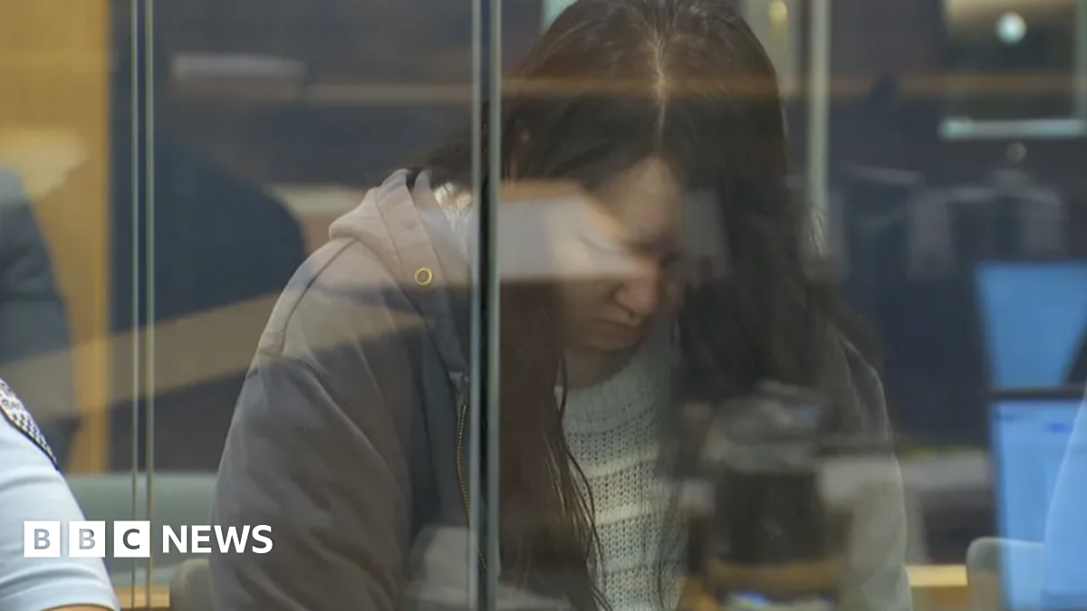 A glum looking woman with her head bowed, seen from behind a glass wall