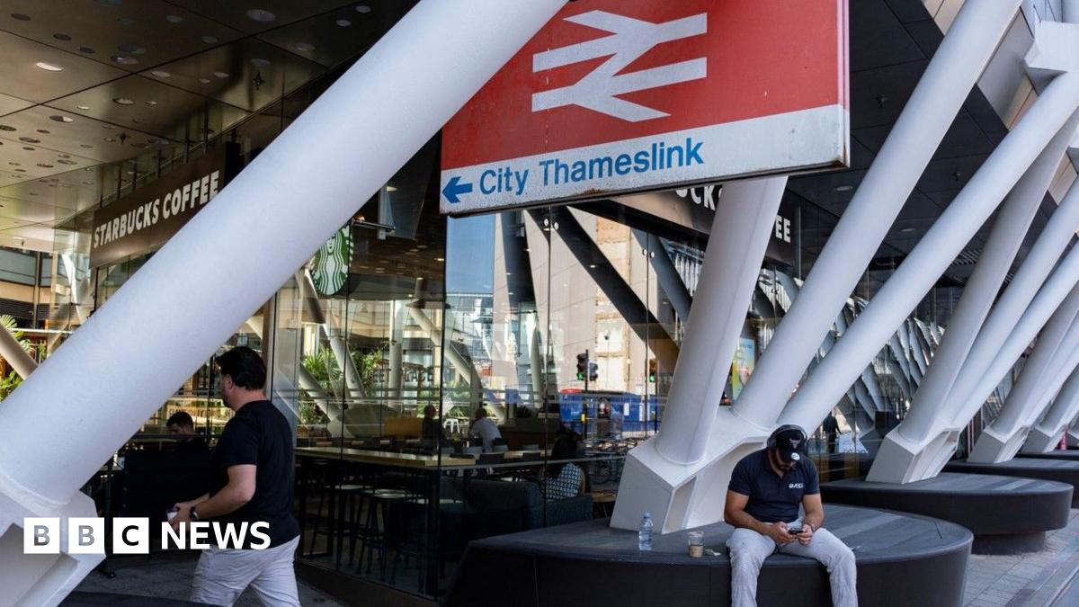 A railway station with a large sign bearing the railway symbol and the words City Thameslink. There are white roof support poles in the foreground, and a Starbucks Coffee in the background. A man is sitting below the sign looking at his mobile phone.