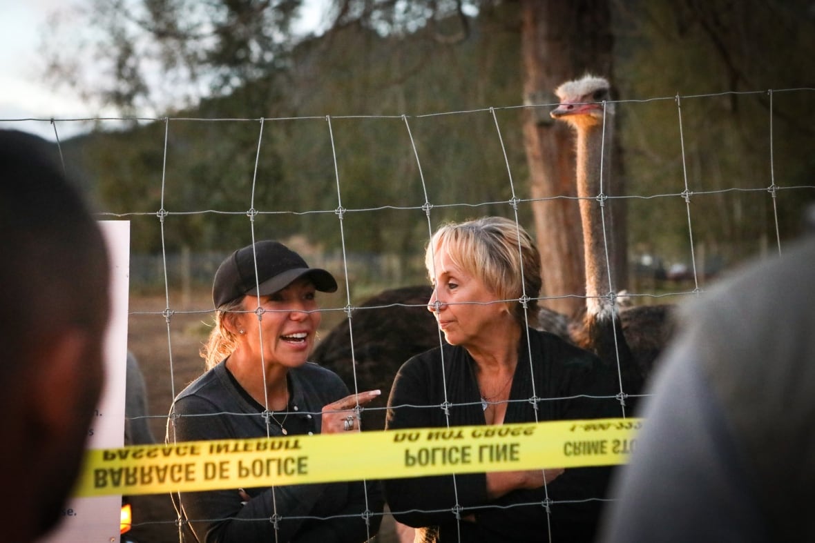 Two women and an ostrich stand behind a wire fence and police tape.