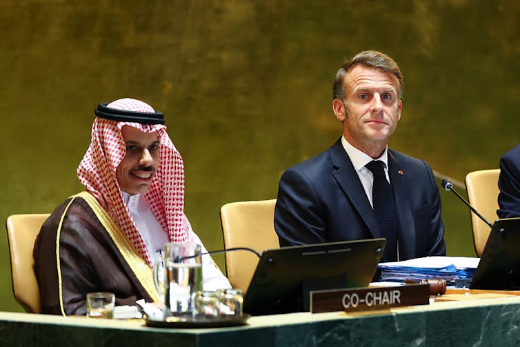 Two men, one in a suit and one wearing a traditional head covering, sit at a table.