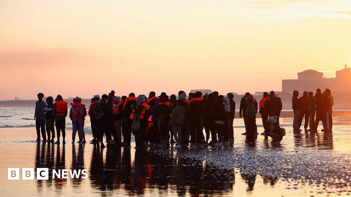 A large group of people standing on a wet beach, many in life jackets, as the sun rises. Their figures are silhouetted and they are reflected on the wet sand. The sea is behind them and the sky is orange and yellow.