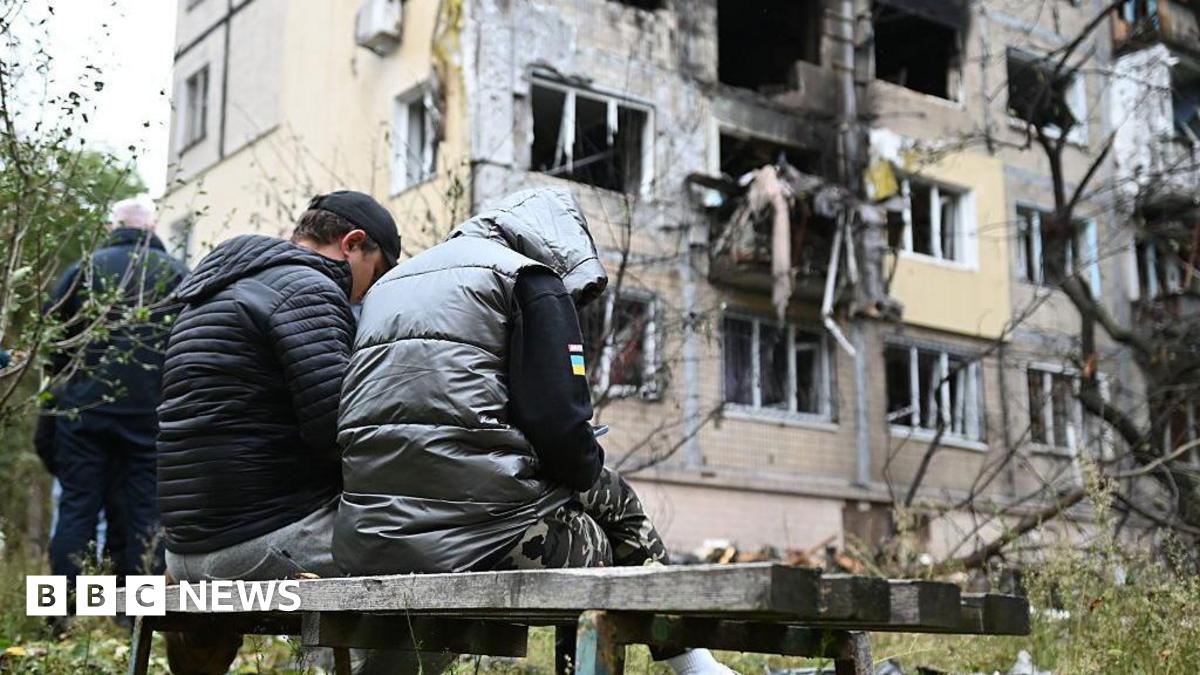 Two people sit on a bench against the backdrop of a destroyed building