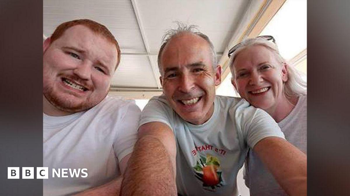 Evan O'Connor and his parents Mark and Louise O'Connor smiling at the camera in a family photo.   Evan has short, ginger hair and a beard.  Mark and Louise both have grey hair.  They are all wearing white t-shirts.