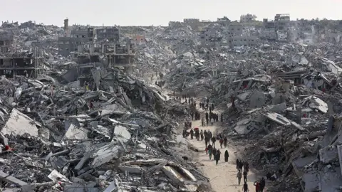AFP via Getty Images A line of people walk down a dirt road, surrounded on each side by tall piles of destroyed buildings. 
