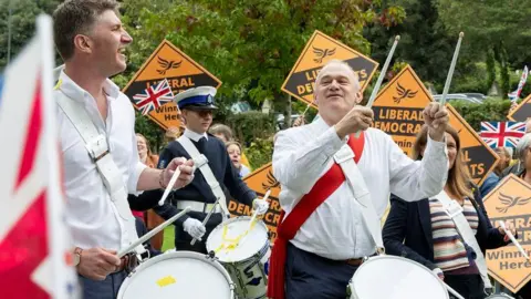 PA Media Ed Davey plays a drum at the head of a marching band as a Union Jack flutters in the breeze next to him