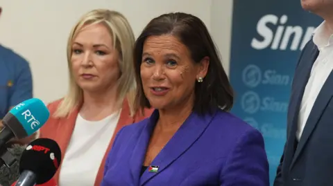PA Media Mary Lou McDonald speaks to microphones. She is wearing a purple blazer and has short dark brown hair. She is stood beside First Minister Michelle O'Neill and TD Pearse Doherty.