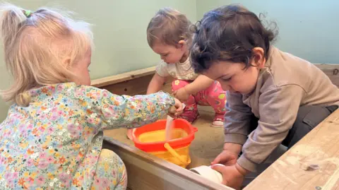 Vanessa Clarke/BBC Three young children play in a wooden sandbox. The one closest to the camera is facing away, pouring sand into a yellow bucket from outside the sandpit. The other two are sat in the sandbox, looking at the sand in their hands below them.