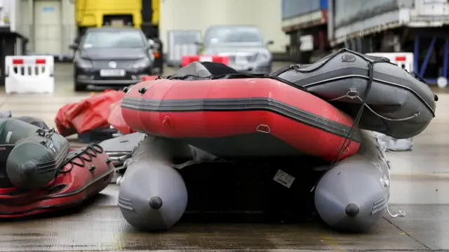 Dinghies piled up on the tarmac outside a port area in the UK. Cars parked in the blurred background