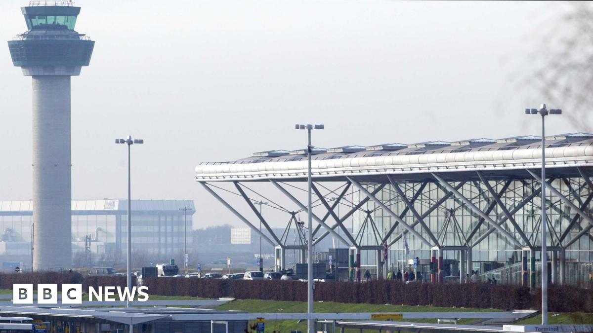 London Stansted Airport on a cloudy day. The airport control tower is to the left and the main terminal buiding, which has a glass facade and a white roof supported by grey pillars.