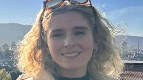 Síofra Harkin Síofra Harkin has long blonde curly hair and is wearing brown and black sunglasses, nestled on her forehead. She is wearing a green top and a beige jacket. 