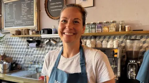 A smiling Annique van Wingerden standing behind the counter at her Windsor cafe with shelves of specialty teas and coffee cups behind her 