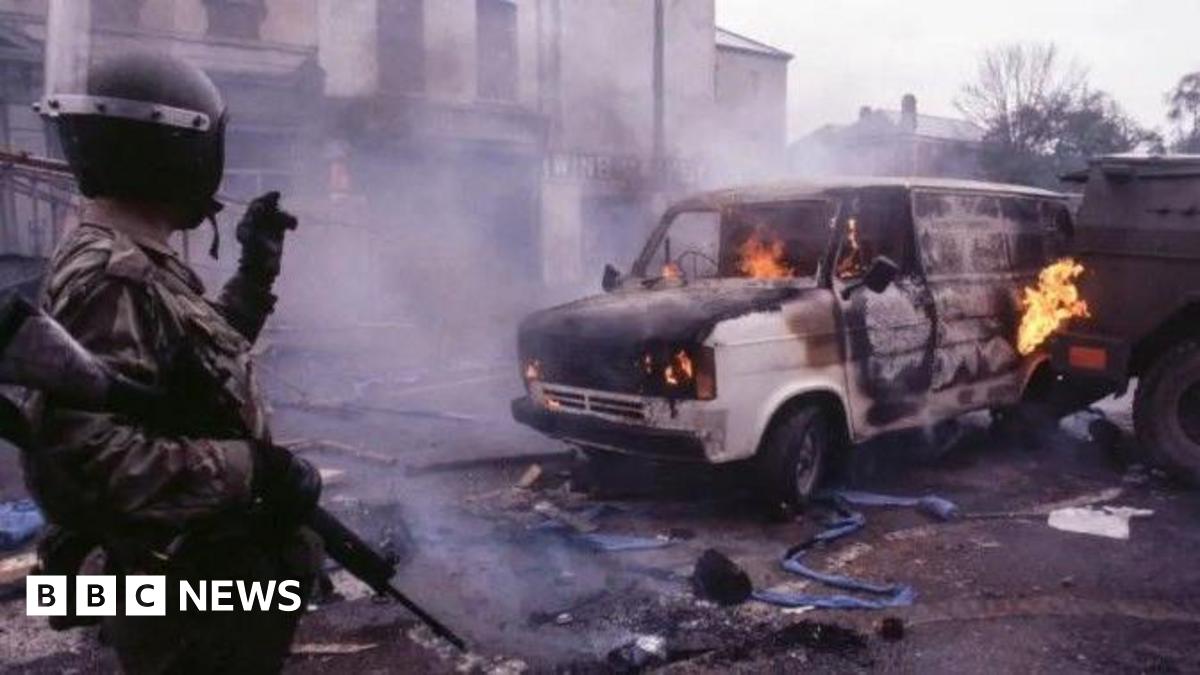 Emmett McConomy is wearing black-rimmed glasses and has a beard. He is dressed in a blue and white polka-dot shirt and is standing outside near a white and brown brick building.