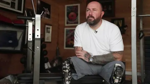 BBC Dave Watson is sitting on a bench in his gym at home. He is wearing a white t-shirt and grey shorts. He has two dark black prosthetic legs covered in stickers. Behind him on the wall are photographs including one of him winning shootout gold medal at the Invictus Games in Sydney.