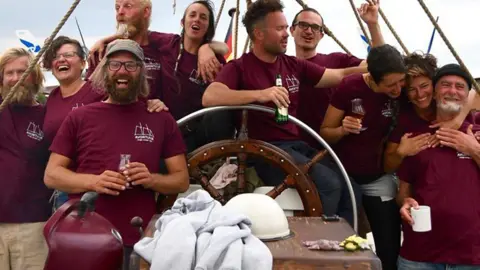 Matthias Berg/Klima Segler The Avontuur crew smile at the camera as they hold up drinks. They are at the helm of the ship and all wearing matching maroon t-shirts. In front of them is a collection of equipment including several helmets and a barbecue