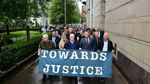 Pacemaker A crowd of people walk down a street holding placards and a banner that reads 'Towards Justice'. There are trees lining the street. The sky is grey.