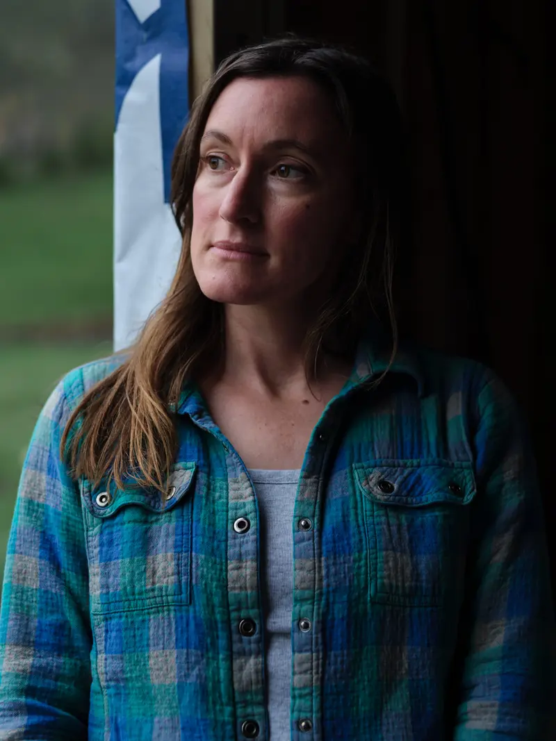 A woman with long brown hair and a blue flannel shirt looks toward light coming in from the left.