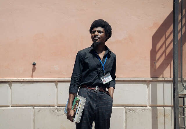 Abubakar stands in front of a wall, holding some books.