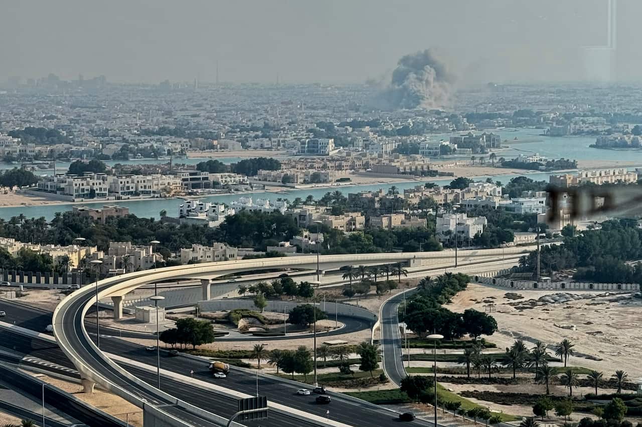 Smoke rises from the site of an airstrike at a distance, with a city visible in the foreground.