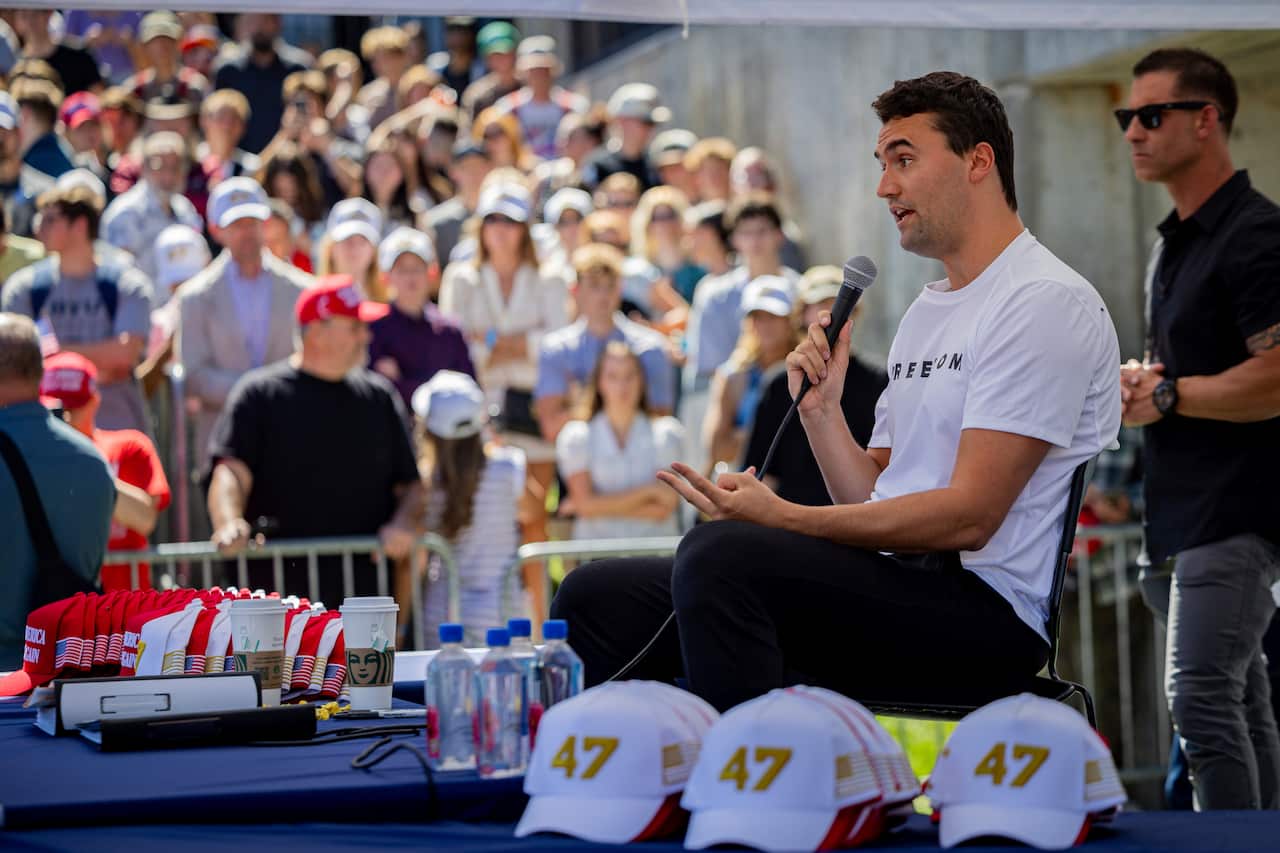 A man in a white round-neck T-shirt sits on a stage, talking into a microphone, as a crowd of young students watches.