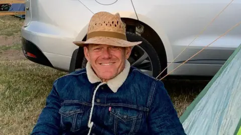 Peter Howard A man sits smiling at the camera. He is wearing a straw hat and demin jacked with a sheeps wool collar. Behind him is the bottom part of a silver car, grass and the edge of a tent. 