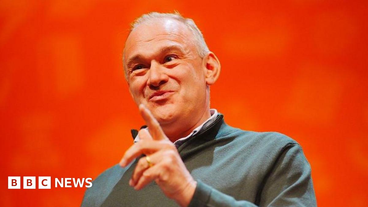 Ed Davey smirks and raises a finger to make a point against a bright orange background, on stage at the Lib Dem conference