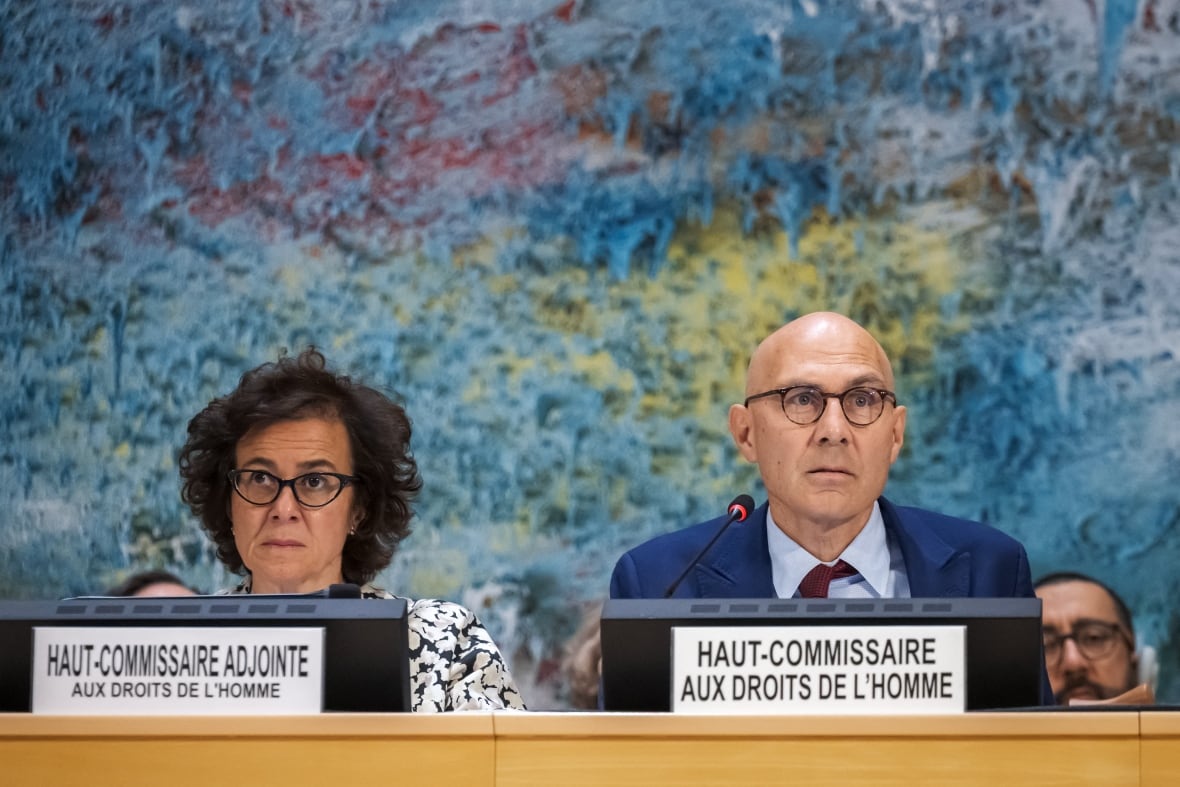 A bald, bespectacled man in a suit and tie, and a bespectacled woman are shown sitting at a long table, with name tags in French in front of them.