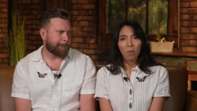 Sky looking over at Amanda who speaks and both look emotional. They sit together on a brown leather sofa in front of a brick work wall and window that looks out onto bushes