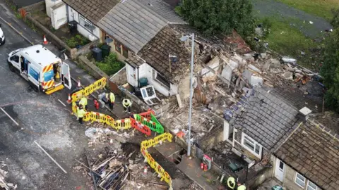 YappApp An aerial shot of a terraced house with extensive explosion damage including a decimated roof and debris all over the road and back garden