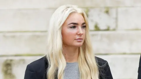 BBC Close up of Megan looking away from the camera. She wears a grey t-shirt and a black blazer. Megan has long blonde hair and black eyelash extensions.