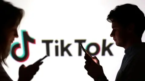 Reuters Teenagers pose for a photo while holding smartphones in front of a TikTok logo