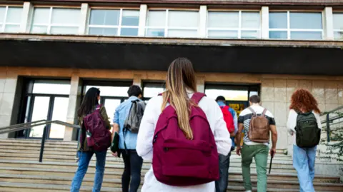 Getty Images A stock image of university students walking up some stairs into a university building. A young woman is at the forefront of the image, she has long blonde hair and is wearing a maroon backpack