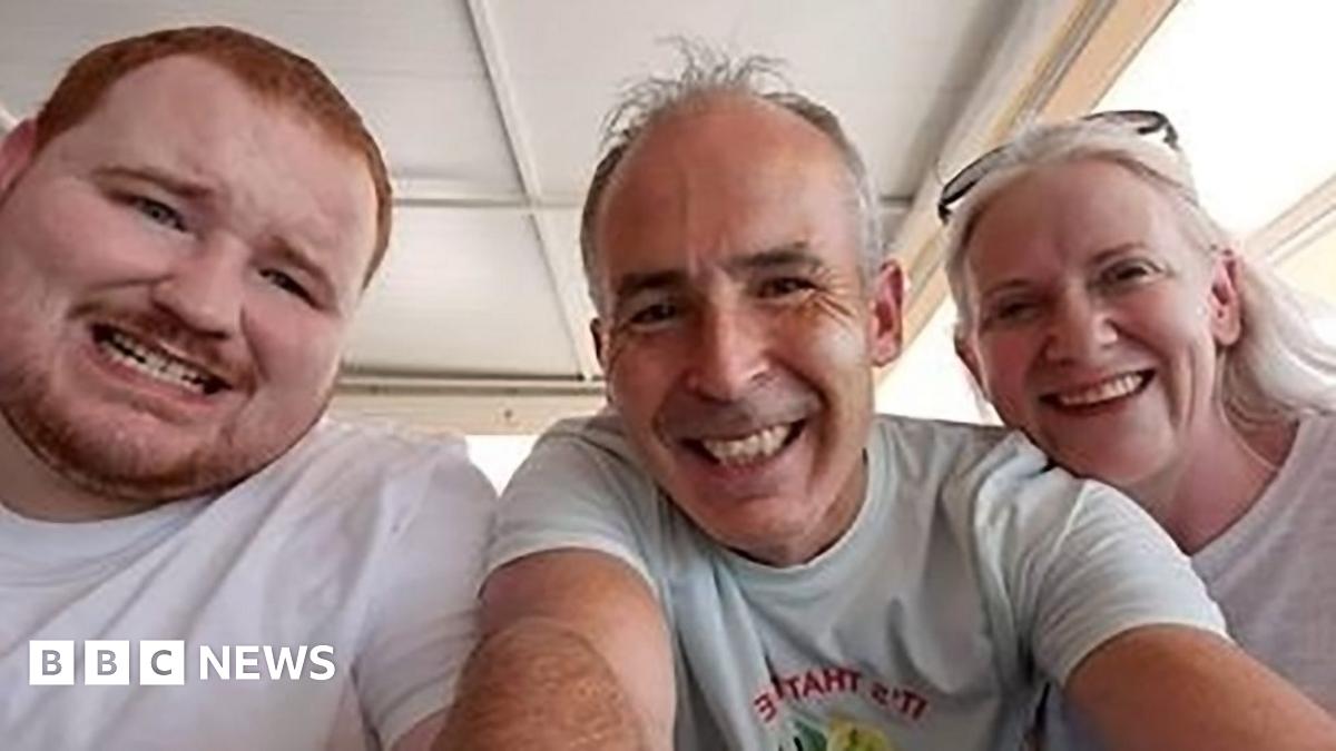 Evan O'Connor and his parents Mark and Louise O'Connor smiling at the camera in a family photo.   Evan has short, ginger hair and a beard.  Mark and Louise both have grey hair.  They are all wearing white t-shirts.