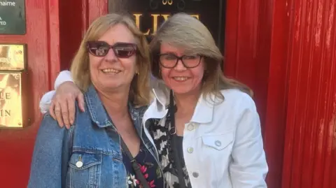 Yvette Greenley Yvette Greenley stands with her arm around her sister Beverley in front of a red door. Both are wearing denim jackets.