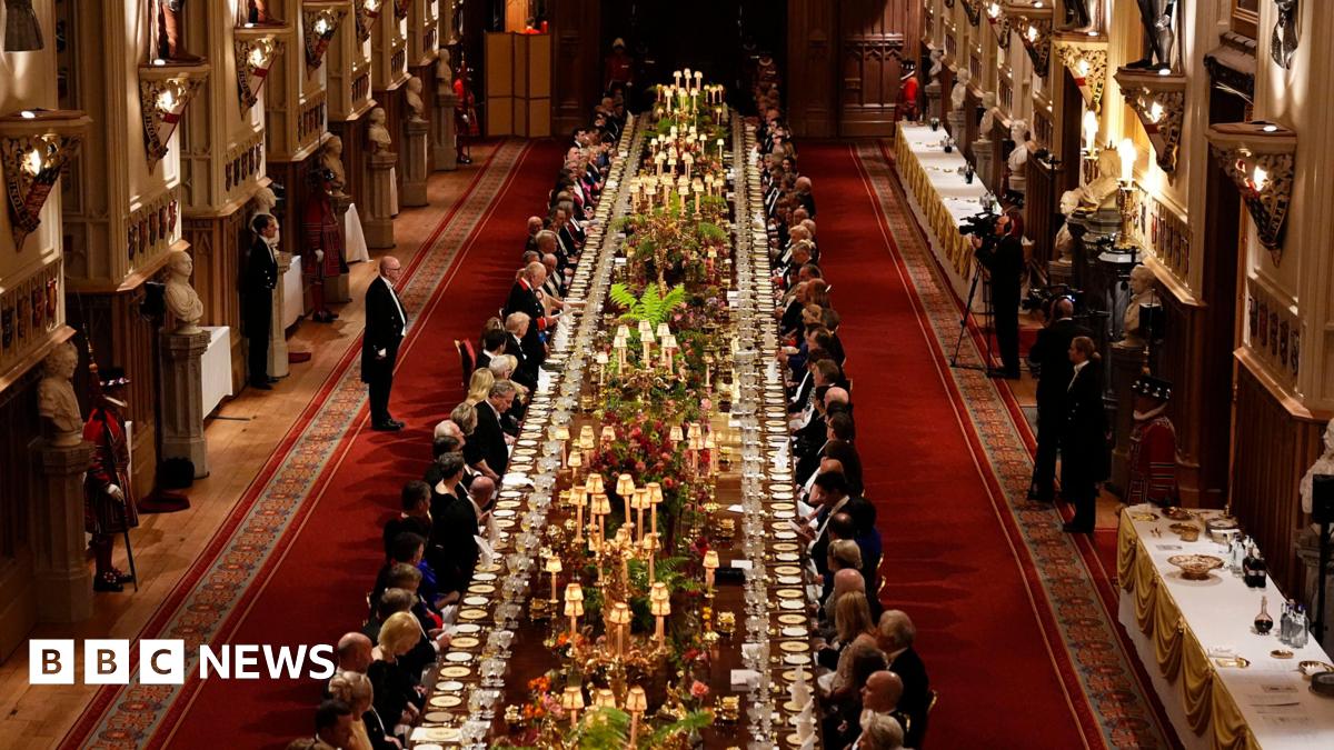 A long dining table with dignitaries seated down either side is seen in a banquet hall, with staff and press against the walls.