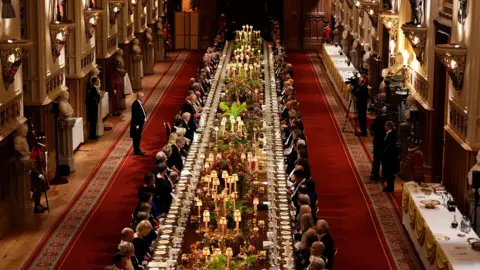 PA A long dining table with dignitaries seated down either side is seen in a banquet hall, with staff and press against the walls.