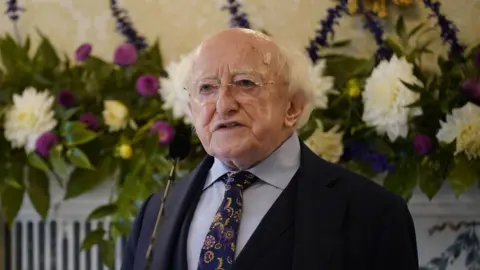 PA Media Michael D Higgins wears a navy suit with a blue shirt and a patterned navy tie. He wears glasses and white hair.